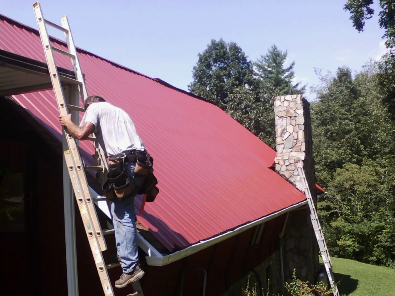 Roofer on a ladder beside a red metal roof and stone chimney — Soffit & Fascia Repair by Roofing Brothers in Big Sandy